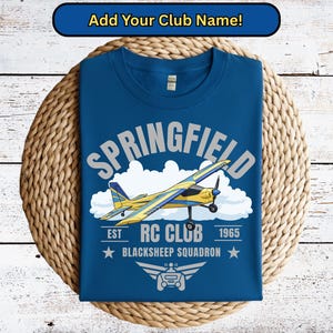 May include: A blue t-shirt with a graphic of a yellow and blue airplane, clouds, and the text "SPRINGFIELD RC CLUB BLACKSHEEP SQUADRON 1965". The shirt is on a woven mat.