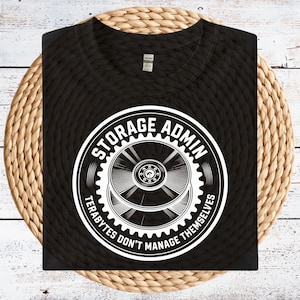 May include: Black t-shirt with a white graphic that reads "STORAGE ADMIN" in a circular design, with the text "TERABYTES DON'T MANAGE THEMSELVES". The graphic features a gear-like design. The shirt is displayed on a woven mat.