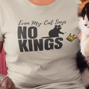 Puede incluir: Una camiseta de color beige claro con un estampado gr&aacute;fico que incluye el texto "Even My Cat Says NO KINGS" en letras gris oscuro. El dise&ntilde;o muestra la silueta de un gato negro alcanzando un peque&ntilde;o icono de corona amarilla a la derecha.