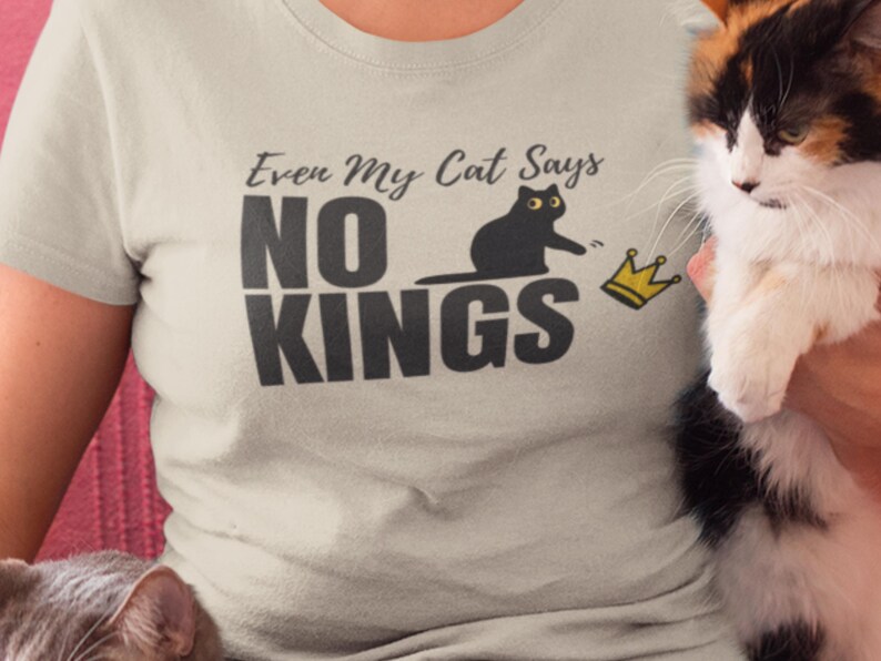 Puede incluir: Una camiseta de color beige claro con un estampado gr&aacute;fico que incluye el texto "Even My Cat Says NO KINGS" en letras gris oscuro. El dise&ntilde;o muestra la silueta de un gato negro alcanzando un peque&ntilde;o icono de corona amarilla a la derecha.