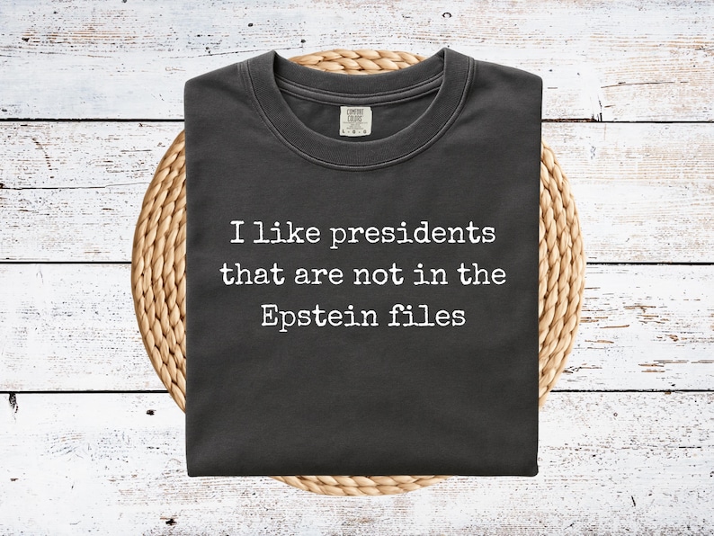 May include: Dark gray t-shirt with the text "I like presidents that are not in the Epstein files" in white. The shirt is folded and rests on a woven, circular placemat, set against a white wooden background.