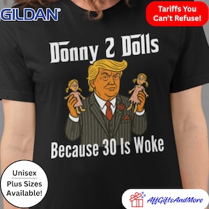 May include: Black t-shirt with a white graphic of a man with blonde hair holding two dolls. The text "Donny 2 Dolls" and "Because 30 Is Woke" is printed on the shirt.