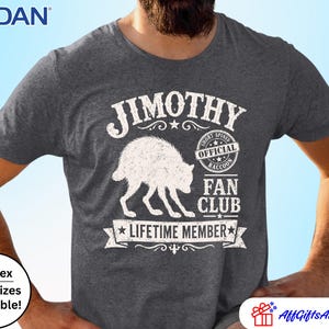 May include: A dark heather grey short-sleeve t-shirt featuring a white graphic of a raccoon. The text reads JIMOTHY, OFFICIAL SHORT-SPINED RACCOON FAN CLUB, and LIFETIME MEMBER. The Gildan brand logo is visible in the top left corner of the image.
