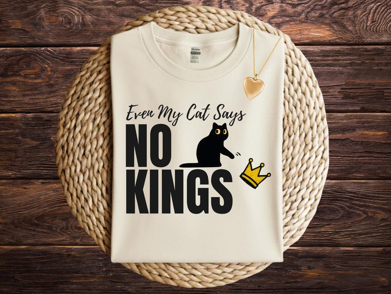 Puede incluir: Una camiseta de color crema con el texto negro "Even My Cat Says NO KINGS" y un gr&aacute;fico de un gato negro apartando una corona amarilla. Un collar con colgante en forma de coraz&oacute;n dorado se sit&uacute;a en la parte superior derecha de la tela.