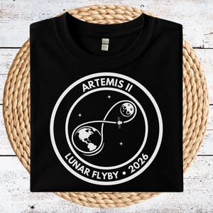 May include: Black t-shirt featuring a white graphic of the Artemis II Lunar Flyby mission. The design includes the Earth, Moon, a spacecraft, and the text "ARTEMIS II LUNAR FLYBY 2026" in a circular arrangement. The shirt is folded.