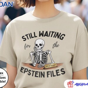 Pode incluir: Camiseta bege com o texto "STILL WAITING for the EPSTEIN FILES" e um gr&aacute;fico de esqueleto. O esqueleto est&aacute; sentado em uma mesa com uma placa de identifica&ccedil;&atilde;o "TRUTH SEEKER". A camiseta &eacute; unissex e est&aacute; dispon&iacute;vel em tamanhos grandes.