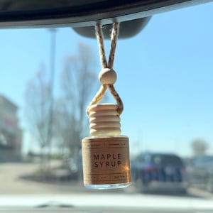 May include: A brown glass bottle with a wooden cap hanging from a rearview mirror. The bottle has a label that reads "Maple Syrup" and is a car air freshener.