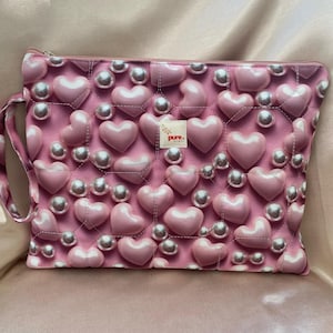 May include: A pink quilted makeup bag with a zipper closure and a strap. The bag is decorated with a pattern of pink hearts and white pearls. The bag has a small logo that says "pure. finds" in white text.