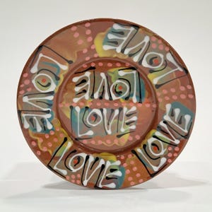 May include: A round ceramic plate with the word "LOVE" repeated in white, black, and red. The plate has a brown base with colorful abstract designs and pink polka dots. A decorative piece.