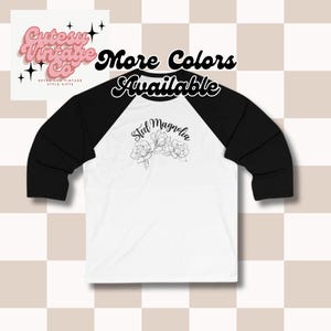 May include: White and black baseball tee shirt with a floral design and the text "Steel Magnolia" on the front. The text "More Colors Available" is on a pink background.