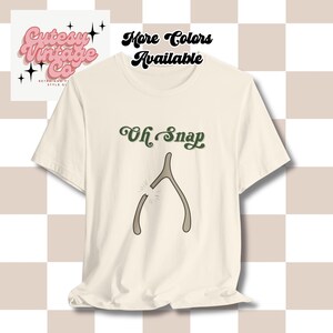May include: Cream-coloured t-shirt with the phrase "Oh Snap" in green script above a broken wishbone graphic. The shirt also has text that says "More Colours Available" and a logo that says "Cutesy Vintage Co."