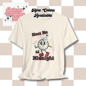 May include: Cream-colored t-shirt featuring a cartoon clock character with a party hat, waving and wearing red shoes. The text "Meet Me at Midnight" is displayed below the clock. The shirt also has the text "More Colors Available".