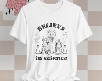May include: White t-shirt featuring a black and white illustration of a cat wearing a lab coat and holding a beaker. The text "BELIEVE in science" is printed below the illustration.