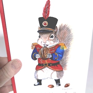 May include: A greeting card featuring a watercolor illustration of a squirrel dressed as a nutcracker soldier. The squirrel wears a red and blue uniform, a black hat with a red feather, and holds a nut. The text "The OG Nutcracker" is at the bottom.
