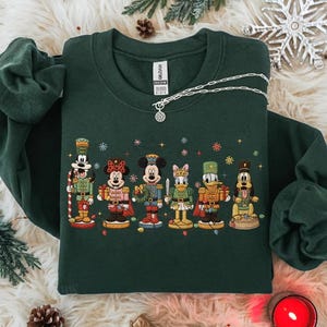 Peut inclure: Sweat-shirt vert foncé orné de personnages de dessins animés en soldats de plomb. Mickey et Minnie Mouse, Donald Duck et Dingo sont représentés. Un collier argenté avec un pendentif est posé sur le vêtement.