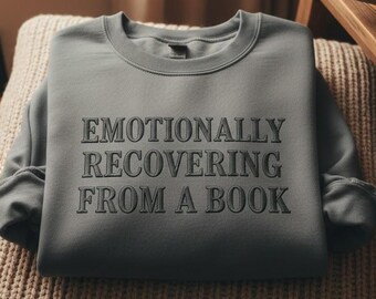 Puede incluir: Una sudadera gris con el texto "EMOTIONALLY RECOVERING FROM A BOOK" en una fuente serif. La sudadera está doblada sobre un cojín de punto beige. Una pequeña mesa de madera con libros y una taza está en el fondo.