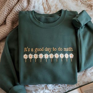 Op de afbeelding: Donkergroene sweatshirt met de geborduurde zin "it's a good day to do math" in gouden letters, samen met een rij madeliefjes. De sweatshirt is opgevouwen op een zacht, crèmekleurig oppervlak.