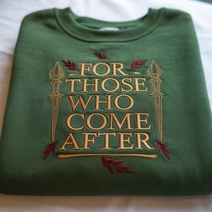 May include: A folded, forest green sweatshirt with embroidered text and decorative elements. The text reads "FOR THOSE WHO COME AFTER" in gold thread, framed by stylized torches and leaves.