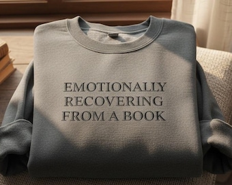 Puede incluir: Una sudadera gris con el texto "EMOTIONALLY RECOVERING FROM A BOOK" en letras negras. La sudadera está doblada sobre un cojín beige. Una vela y libros son visibles en el fondo.