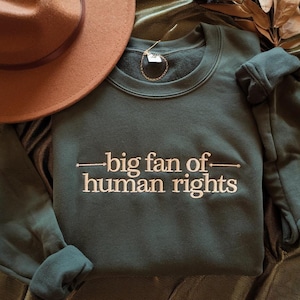 Könnte beinhalten: Ein dunkelgrünes Sweatshirt mit dem weißen Text "big fan of human rights". Ein brauner Hut ist in der oberen linken Ecke zu sehen. Das Sweatshirt ist gefaltet, und das Bild ist von oben aufgenommen.