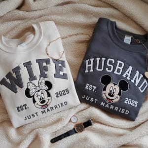 Puede incluir: Dos sudaderas a juego, una crema y otra gris carb&oacute;n, con las palabras "WIFE" y "HUSBAND" en negrita. Cada una presenta un dise&ntilde;o de Mickey Mouse y el texto "EST. JUST MARRIED 2025". Tambi&eacute;n se ven collares dorados y un reloj.