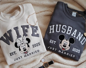 May include: Two matching sweatshirts, one cream and one charcoal gray, with "WIFE" and "HUSBAND" in bold letters. Each features a Mickey Mouse design and the text "EST. JUST MARRIED 2025". Gold necklaces and a watch are also visible.