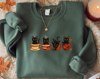 Puede incluir: Sudadera verde con un diseño bordado de gatos negros en tazas de té con detalles otoñales. El diseño incluye calabazas y hojas. También se ve un collar dorado y una vela encendida.