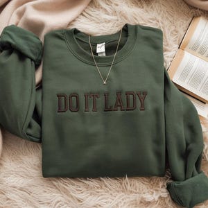 Könnte beinhalten: Ein waldgrünes Sweatshirt mit Rundhalsausschnitt und dem gestickten Text DO IT LADY in dunkelbraunen Blockbuchstaben auf der Brust. Das Kleidungsstück hat lange Ärmel mit gerippten Bündchen und einen gerippten Ausschnitt sowie eine goldfarbene Kette.