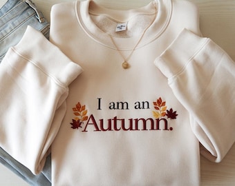 May include: Cream-colored sweatshirt with the embroidered text "I am an Autumn" in a burgundy color, accompanied by fall leaf designs. A gold necklace and blue jeans are also visible.