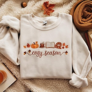 Könnte beinhalten: Cremefarbenes Sweatshirt mit gesticktem Herbstdesign. Das Design umfasst einen Kürbis, eine Tasse, ein offenes Buch und ein Gebäck, mit dem Schriftzug "cozy season" in Schreibschrift. Das Sweatshirt ist von herbstlichen Accessoires umgeben.