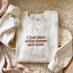 May include: A cream-coloured sweatshirt embroidered with the phrase "I just want some cheese and quiet." A gold necklace with a small pendant is on the shirt. The sweatshirt has long sleeves with ribbed cuffs.