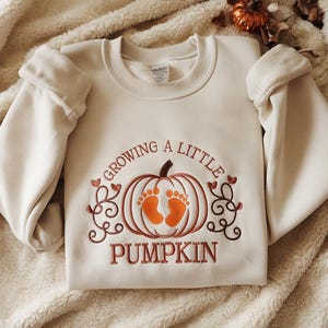 May include: Cream-coloured sweatshirt with the embroidered text "Growing a Little Pumpkin." The design features a pumpkin with orange baby footprints inside. The sweatshirt has long sleeves and a crew neck.