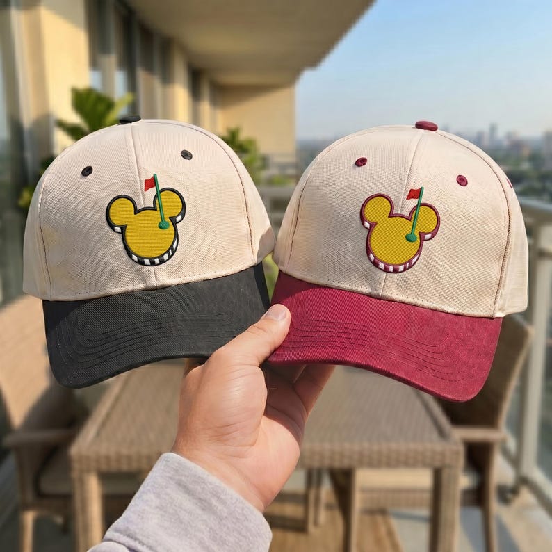 May include: Two baseball caps with a golf-themed design. The caps have a cream-colored crown, a black and burgundy brim, and a yellow golf ball graphic. The caps are held up by a person's hand.