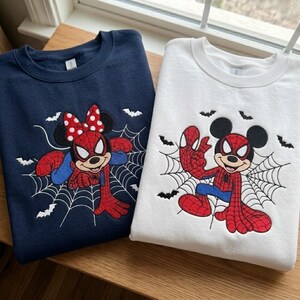 May include: A navy blue sweatshirt and a white sweatshirt, each featuring an embroidered graphic of a cartoon character dressed as a superhero. The navy sweatshirt shows a character with a red polka-dot bow, while the white sweatshirt shows a character with round ears, both positioned against a black spiderweb and small bat designs.