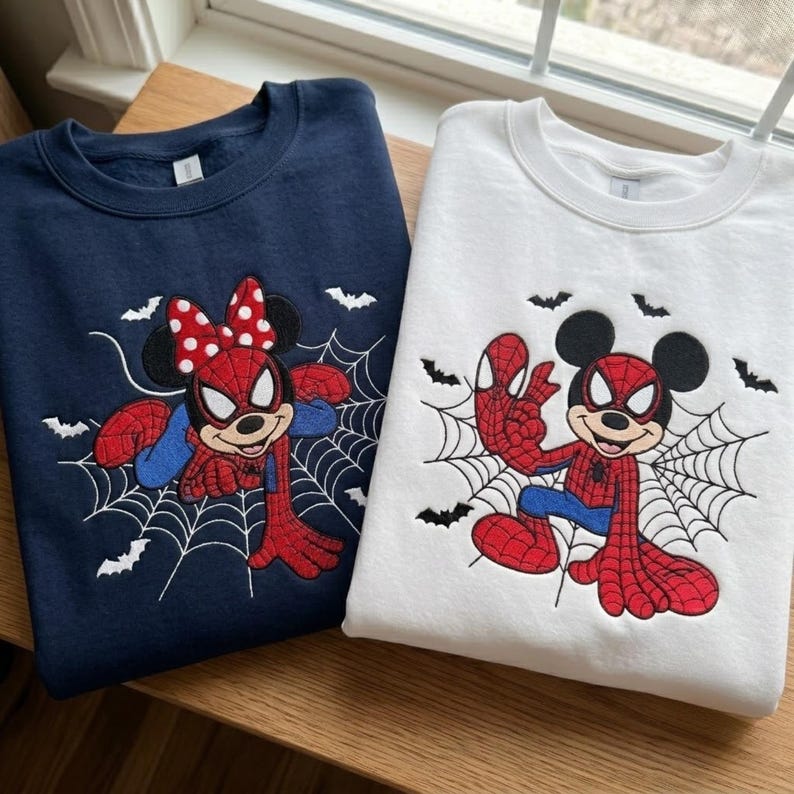 May include: A navy blue sweatshirt and a white sweatshirt, each featuring an embroidered graphic of a cartoon character dressed as a superhero. The navy sweatshirt shows a character with a red polka-dot bow, while the white sweatshirt shows a character with round ears, both positioned against a black spiderweb and small bat designs.