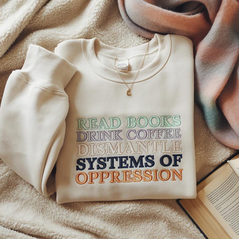 May include: Cream-colored sweatshirt with embroidered text: "READ BOOKS DRINK COFFEE DISMANTLE SYSTEMS OF OPPRESSION." A gold necklace with a small pendant rests on the shirt. The sweatshirt is folded on a textured surface with a blanket and open book in the background.