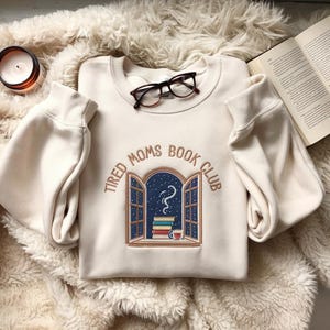 Może przedstawiać: Kremowa bluza z napisem "Tired Moms Book Club" nad haftowaną sceną okna z książkami, filiżanką i gwiaździstą nocą. Okulary spoczywają na bluzie.