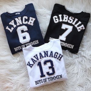 May include: Three white sweatshirts with navy blue and gold lettering. The sweatshirts have the names "Lynch", "Gibsile", and "Kavanagh" with corresponding numbers 6, 7, and 13. Each sweatshirt has the text "Boys of Tommen" below the name and number.