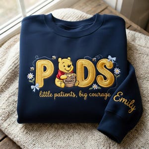 May include: Navy blue sweatshirt with gold embroidered text and a Winnie the Pooh design. The word "Peds" is featured, with Winnie the Pooh holding a honey pot. The sleeve has the name "Emily" embroidered in gold. The text "little patients, big courage" is also included.