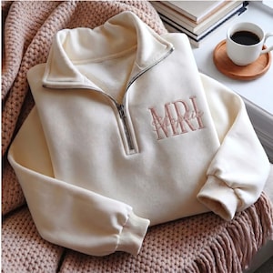 Può includere: Una felpa color crema con zip a un quarto, collo alto e cerniera in metallo argentato. Sul petto è presente un ricamo color rosa antico con la scritta MRI e il nome Sophia scritto in corsivo sovrapposto alle lettere.