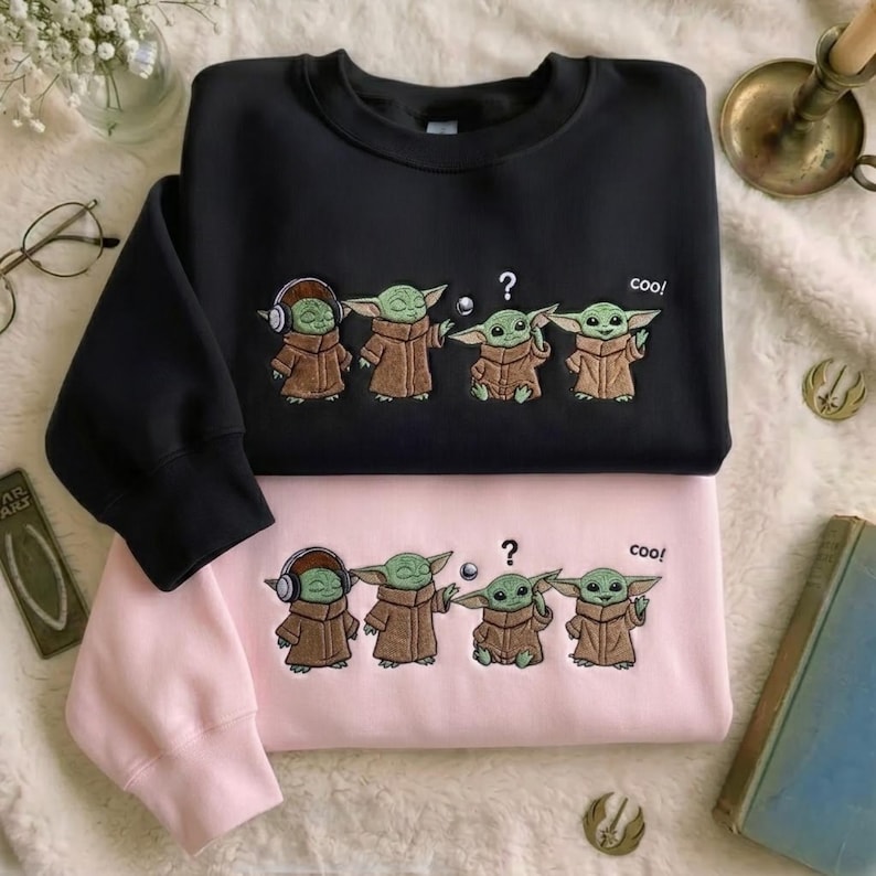 Puede incluir: Una sudadera negra y una sudadera rosa claro, cada una con un gr&aacute;fico bordado de cuatro criaturas verdes con t&uacute;nicas marrones. El dise&ntilde;o incluye un signo de interrogaci&oacute;n, una peque&ntilde;a esfera y el texto coo! sobre las figuras.