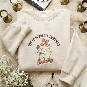 Op de afbeelding: Crèmekleurige sweatshirt met de geborduurde tekst "OFF TO REGULATE EMOTIONS". Het ontwerp toont een gans op een roze scooter die een koffiebeker vasthoudt. De gans heeft een roze strik en is omringd door sterren.