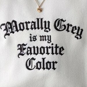 May include: Cream-colored sweatshirt with the embroidered phrase "Morally Grey is my Favorite Color" in black gothic lettering. A gold-colored necklace with a small pendant is visible at the top. The fabric has a soft texture.