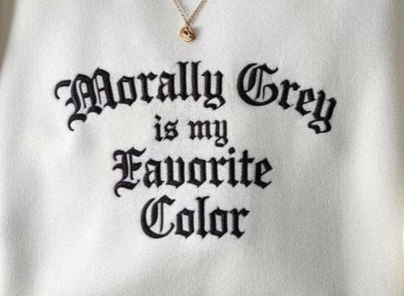 May include: Cream-colored sweatshirt with the embroidered phrase "Morally Grey is my Favorite Color" in black gothic lettering. A gold-colored necklace with a small pendant is visible at the top. The fabric has a soft texture.