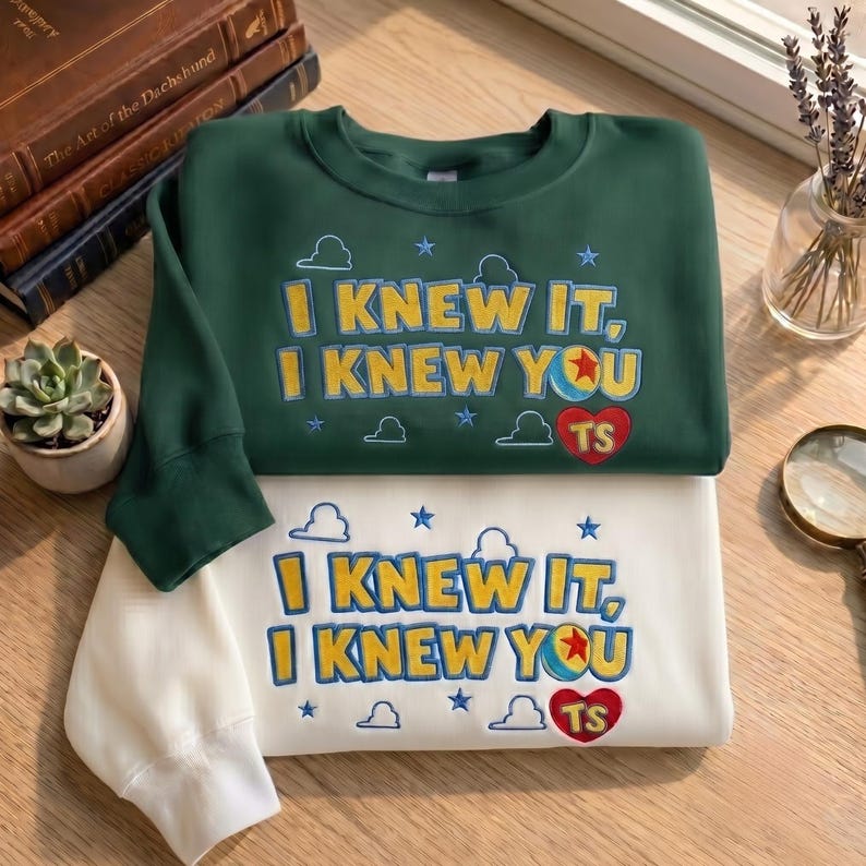 May include: A dark green sweatshirt and a white sweatshirt, each featuring embroidered text that reads I KNEW IT, I KNEW YOU, accompanied by a red heart with the letters TS, a colorful ball with a red star, and small blue stars and clouds.