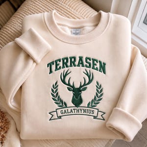 Puede incluir: Una sudadera de cuello redondo color crema con bordado verde oscuro en el pecho. El diseño incluye el texto TERRASEN arqueado sobre un emblema central de cabeza de ciervo flanqueado por ramas de laurel, con el texto GALATHYNIUS en una cinta.