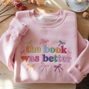 May include: A light pink sweatshirt with the embroidered text "the book was better" in rainbow colors. The sweatshirt has a crew neck and rolled-up sleeves. Decorative bows are embroidered around the text.