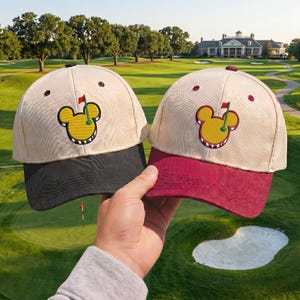 May include: Two beige golf hats with black and maroon brims. Each hat features a yellow Mickey Mouse-shaped golf ball design with a red flag. The hats are held up against a golf course background.