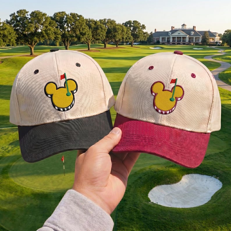May include: Two beige golf hats with black and maroon brims. Each hat features a yellow Mickey Mouse-shaped golf ball design with a red flag. The hats are held up against a golf course background.
