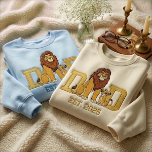 May include: Two crewneck sweatshirts in light blue and cream, each featuring an embroidered graphic of a lion and cub inside the letters of the word DAD. The cream sweatshirt includes the embroidered text EST. 2025 below the graphic design.